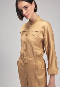 Vestido camisero de satén en tono marrón dorado, con cierre de botones en la parte delantera, cuello y bolsillo en el pecho. Presenta una cintura fruncida con un cinturón redondo y bolsillos laterales.