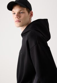 Melnā kapuces džemperis ar teksturētu virsmu, ar izšūtu logotipu uz krūts un brīvu piegriezumu. Modelis redzams no sāna.