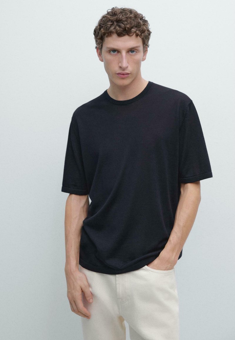 Jeune homme aux cheveux bouclés portant une chemise noire à manches courtes et un pantalon beige clair, debout devant un fond clair uni.