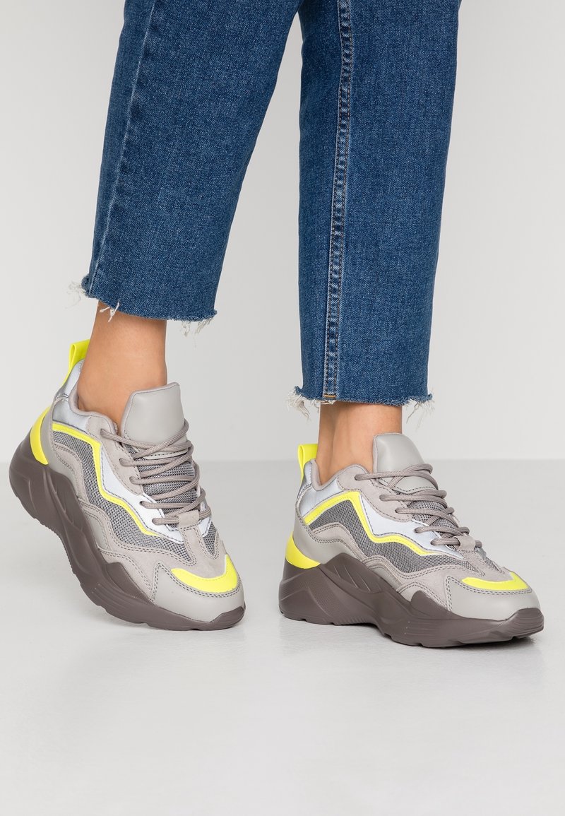 Pieds portant des baskets épaisses grises et jaunes associées à un jean bleu raccourci sur un fond clair uni.