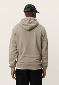 Μπεζ φούτερ με κουκούλα και μαλακή υφή, με τσέπη καγκουρό και ριμπ μανσέτες. Συνδυασμένο με μαύρο καπελάκι μπέιζμπολ με λουλουδάτο σχέδιο.