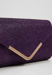 Pochette texturée violette avec un accent métallique doré en forme de V sur le rabat avant, sur fond gris clair.