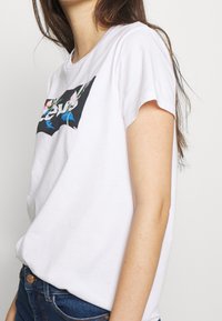 Valkoinen lyhytniminen t-paita, jossa on suorakulmainen musta grafiikka kukkakuviolla ja "Levi's"-teksti rohkeilla valkoisilla kirjaimilla esillä.