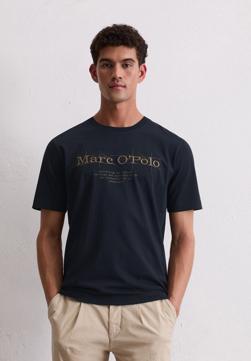 Navyblaue Baumwoll-T-Shirt mit kurzen Ärmeln. Hat auffälligen goldenen Text mit der Aufschrift "Marc O'Polo" und einer Umweltnachricht darunter.
