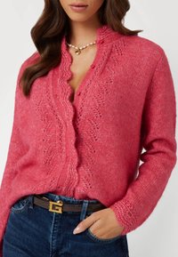 Roze gebreide cardigan met gekartelde randen, decoratieve stiksels en knoopsluiting; gecombineerd met donkerblauwe jeans en een leren bruine riem.
