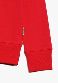 Manga de sudadera roja con puño acanalado. Textura de tela suave, presenta una pequeña etiqueta de marca cerca de la costura.