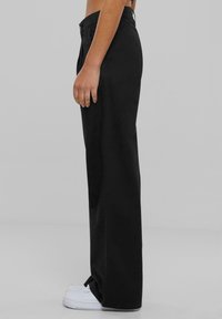 Persona con pantalones negros sueltos y zapatillas blancas, vista de perfil sobre un fondo gris liso.