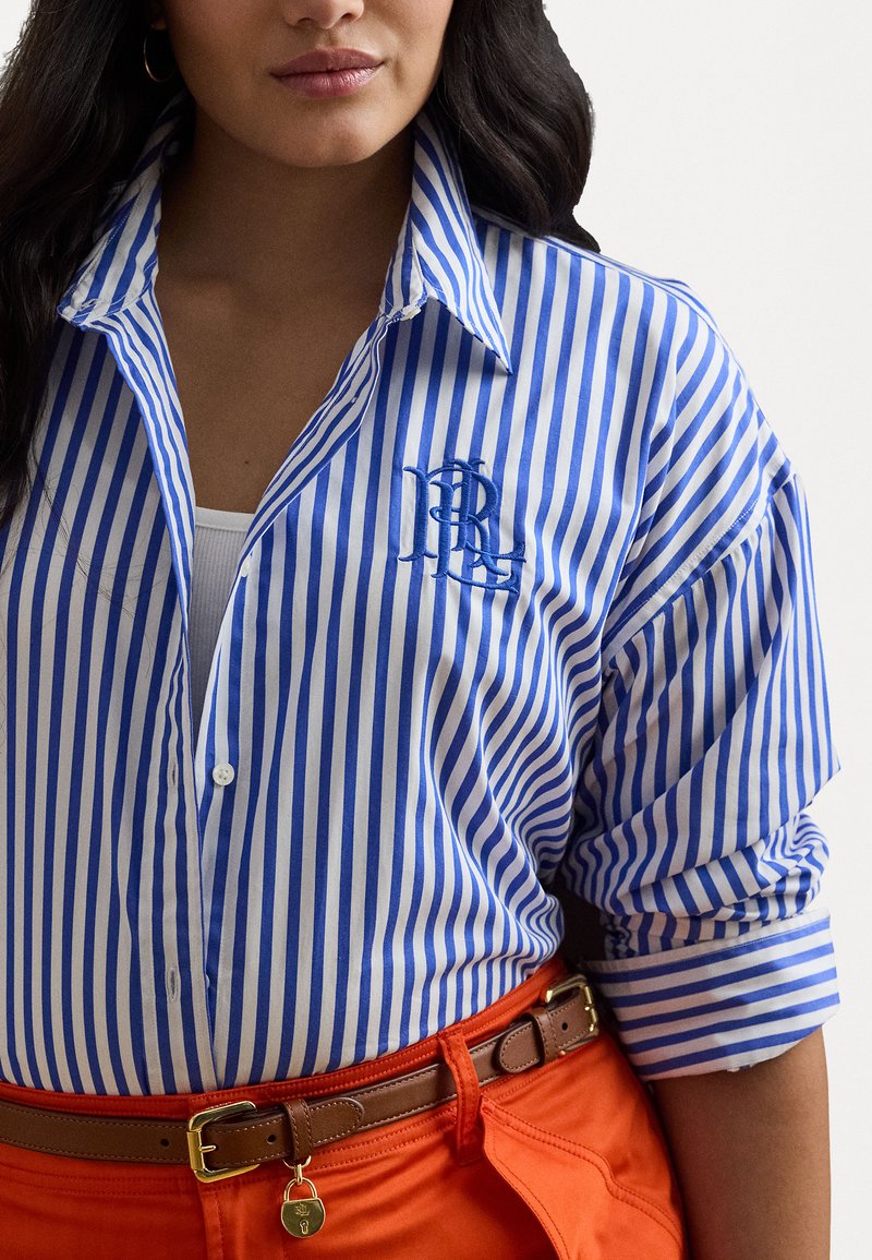 Camisa de botones a rayas azules y blancas con un logo bordado, combinada con pantalones naranjas y un cinturón marrón con un detalle de herrajes dorados.