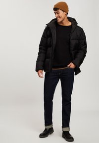 Czarna kurtka puchowa z kapturem, z zapięciem na zamek i przednią kieszenią. Noszona na czarnym swetrze, zestawiona z ciemnoniebieskimi dżinsami i czarnymi botkami.