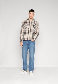 Camisa xadrez em bege e castanho com dois bolsos no peito, combinada com jeans azul claro e ténis brancos com detalhes em castanho.