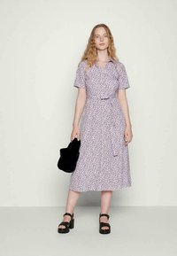 Mulher com cabelo longo e ondulado, vestindo um vestido midi floral em lavanda, segurando uma bolsa preta e sandálias de plataforma pretas, de pé contra um fundo simples.