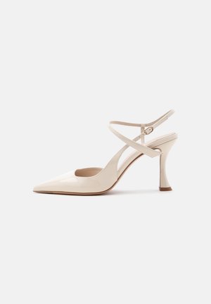 Chaussure à talons hauts en cuir verni beige, bout pointu et fines brides croisées à la cheville avec fermeture par boucle sur fond blanc.