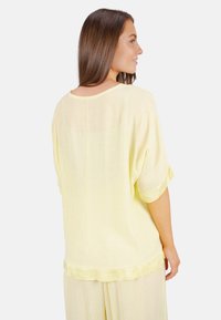 Helle gelbe, lockere Bluse aus strukturiertem Stoff mit rundem Ausschnitt und hochgerollten Ärmeln. Die Rückansicht zeigt dezente Nahtdetails.