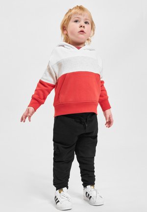 Un tout-petit aux cheveux blonds portant un sweat à capuche rouge et blanc, un pantalon noir et des baskets Adidas blanches, se tenant contre un fond uni.