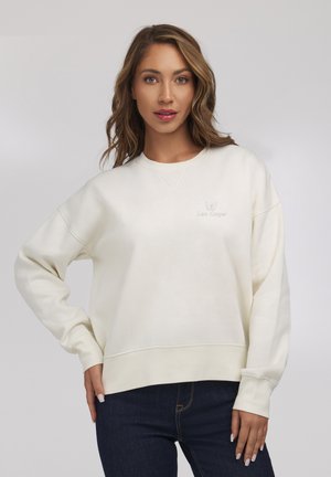 Kobieta z falującymi włosami, ubrana w kremową bluzę z małym logo motyla i ciemne dżinsy, stojąca na tle jednolitego jasnego tła.