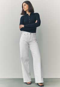 Čierny priliehavý top s dlhým rukávom v kombinácii s bielymi širokými nohavicami s vysokým pásom. Nohavice majú predné vrecká a zapínanie na gombík. Čierne sandále so zápätiami.