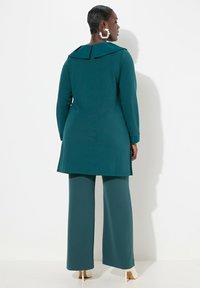 Tunica teal con un ampio colletto, maniche lunghe e fessure laterali abbinata a pantaloni con gamba larga. Tessuto morbido con un design aderente.