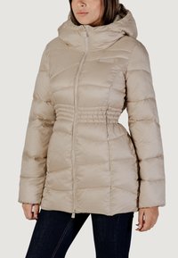 Donna che indossa un giubbotto imbottito beige con cappuccio, zip frontale e vita elasticizzata, abbinato a jeans blu scuro.