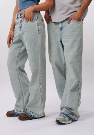 Dvaja ľudia oblečení v svetlomodrých širokých džínsach s ošúchanými lemami, jeden má na nohách hnedé topánky, druhý sivé tenisky, s rukami vo vreckách.