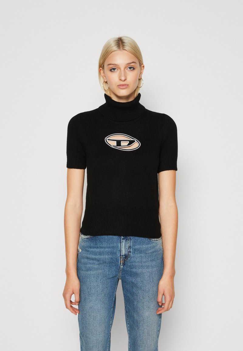 Schwarzer kurzärmeliger Rollkragenpullover mit gerippter Textur, ausgestattet mit einem beigefarbenen und schwarzen ovalen Grafiklogo in der Mitte. Kombiniert mit blauer Jeans.