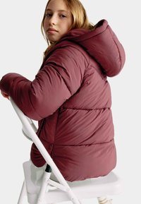 Burgundy vadderad jacka med huva, med ett kviltat mönster, mjuk textur och en figursydd passform, bärs när personen sitter på en vit stol.
