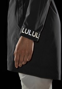 Svart vattentät kappafunktion med en logotyp på ärmen. Slät textur med en kroppsnära design och en synlig hand som håller plagget.