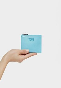 Tarjetero de cuero azul con cierre de cremallera, logo "TOUS" en relieve en la parte frontal, textura suave, forma rectangular, sostenido en una mano.