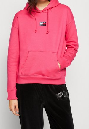 Osoba nosząca jasnoróżową bluzę z kapturem Tommy Jeans z przednią kieszenią oraz czarne spodnie z białym napisem na udzie.