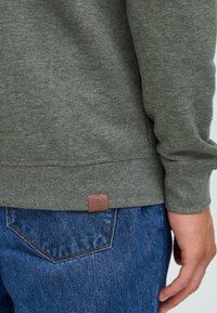 Sweatshirt en coton vert avec des poignets et un ourlet côtelés, présente un patch en cuir marron en bas à gauche, associé à un jean en denim bleu.