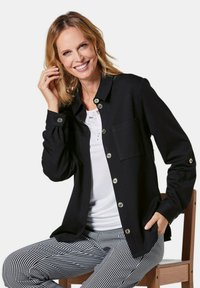 Zwarte button-up jas met lange mouwen, met een borstzak en zilveren hardware, gedragen over een witte top en geruite broek.