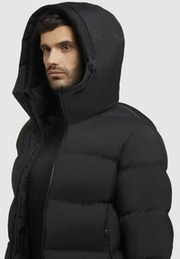 Sort pufferjakke med høj krave og oversize hætte. Har quiltede sektioner og lynlås foran. Glat, blød tekstur.