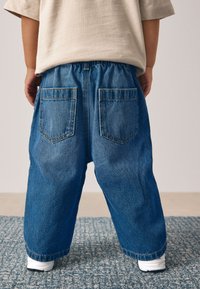 Blugi din denim cu croială lejeră, culoare albastru deschis, talie elastică și două buzunare în spate. Purtate cu pantofi sport albi.