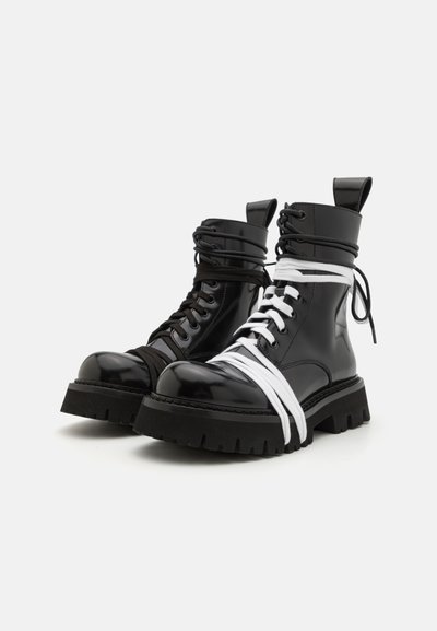 Botas de tobillo de charol negro con punta redonda, suela gruesa negra y cordones blancos y negros en contraste entrelazados alrededor de las cañas.
