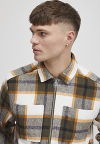 Flannel zip-up jacket with a plaid pattern in shades of olive, orange, gray, and white; features a collar and two front pockets.