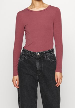 Osoba w dopasowanej, pudrowo-różowej bluzce z długim rękawem, wpuszczonej w czarne, wyprane dżinsy z wysokim stanem i zapięciem na guziki z przodu.