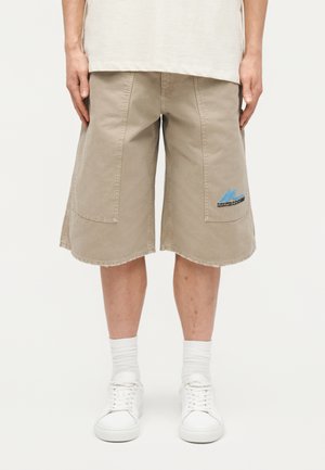 Personne portant un short cargo beige mi-cuisses avec un logo bleu sur la poche droite, des chaussettes blanches et des baskets blanches, debout devant un fond uni.