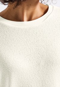 Pull crème clair avec une texture lâche, encolure ronde et poignets côtelés. Motif légèrement ajouré qui ajoute un intérêt visuel.