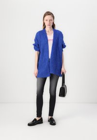 Blå, myk cardigan med løs passform og åpen front, brukt over en lys rosa grafisk t-skjorte. Kombinert med mørke jeans og svarte loafers.