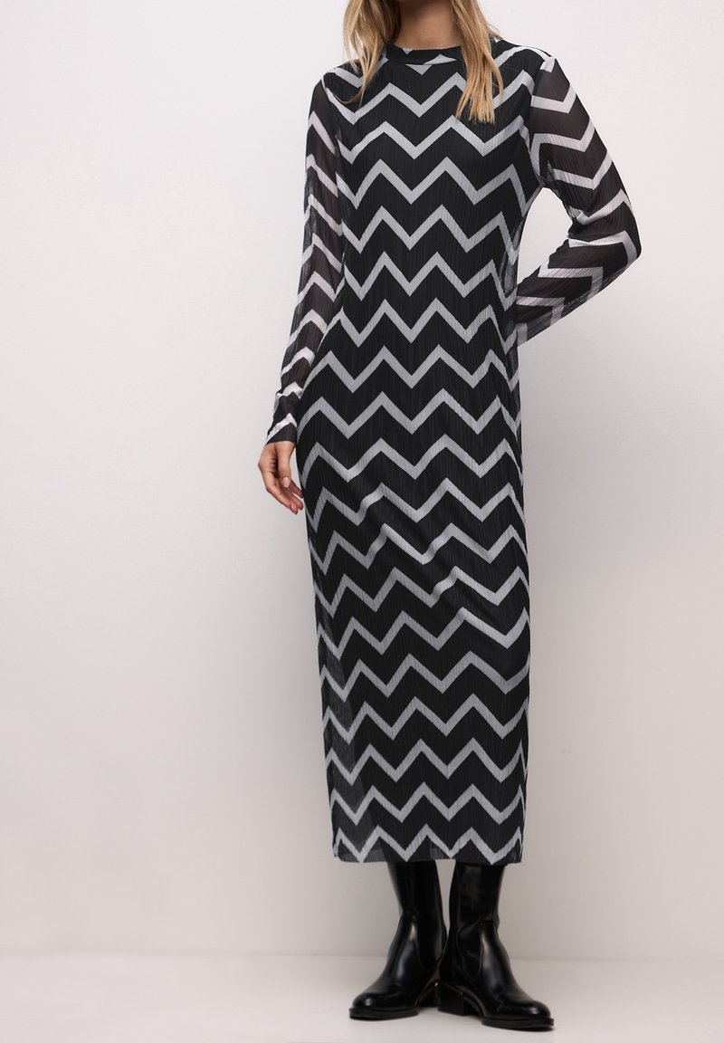 Femme portant une robe midi noire à manches longues avec un motif zigzag blanc et des bottines noires, debout devant un fond clair uni.