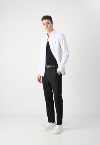 Camicia bianca a maniche lunghe sopra una canotta nera, pantaloni neri e sneakers bianche. Dettagli notevoli includono un logo sulla camicia e una cintura con texture.