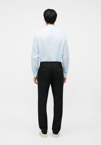 Άνδρας που στέκεται με την πλάτη, φοράει ανοιχτό μπλε μακρυμάνικο πουκάμισο, μαύρο παντελόνι και λευκά αθλητικά παπούτσια, μπροστά σε απλό φόντο.