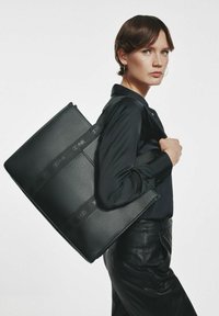 Kobieta z krótkimi brązowymi włosami, ubrana w czarną koszulę i skórzane spodnie, nosi dużą czarną torbę teksturalną na ramieniu, na tle białego tła.