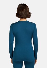 Woman with short brown hair wearing a fitted long-sleeve teal top and matching pants, shown from the back on a white background.