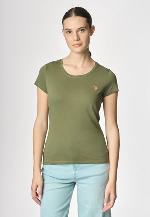 Modella che indossa una maglietta verde con un piccolo logo, abbinata a jeans azzurri chiari, si trova davanti a uno sfondo neutro, guardando direttamente verso la fotocamera.