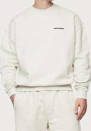 Mand iført off-white sweatshirt og matchende bukser med "PEGADOR" logo på venstre bryst, hænder delvist i lommer.