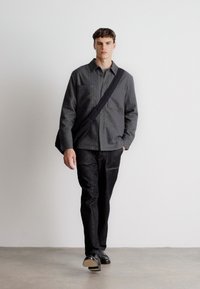 Szürke gombos ing, két mellzsebbel, sötét nadrággal párosítva. A modell egy minimalistának mondható fekete válltáskát hord. Fekete cipők.