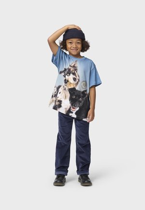 Kind lächelt, trägt eine blaue Mütze, übergroßes T-Shirt mit Hundemotiv, dunkle Hose und schwarze Schuhe, steht vor einem einfarbigen hellen Hintergrund.