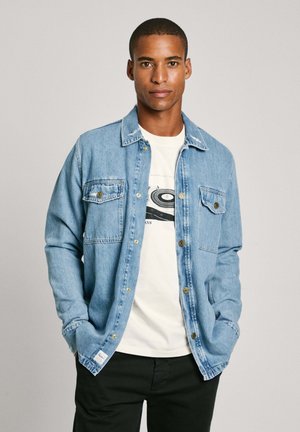 Ljust blå denimskjorta med knäppning fram, två bröstfickor och en avslappnad passform. Bärs över en vit t-shirt och matchas med svarta byxor.