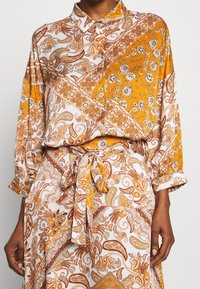 Chemise à manches longues blanche et orange, avec imprimé paisley, col boutonné et détail de ceinture à nouer, coupe ample et texture de tissu douce.