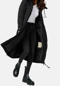 Lang sort dunjakke med hætte, lynlåslukning og indvendige lommer, kombineret med tætsiddende sorte jeans og chunky sorte ankelstøvler.